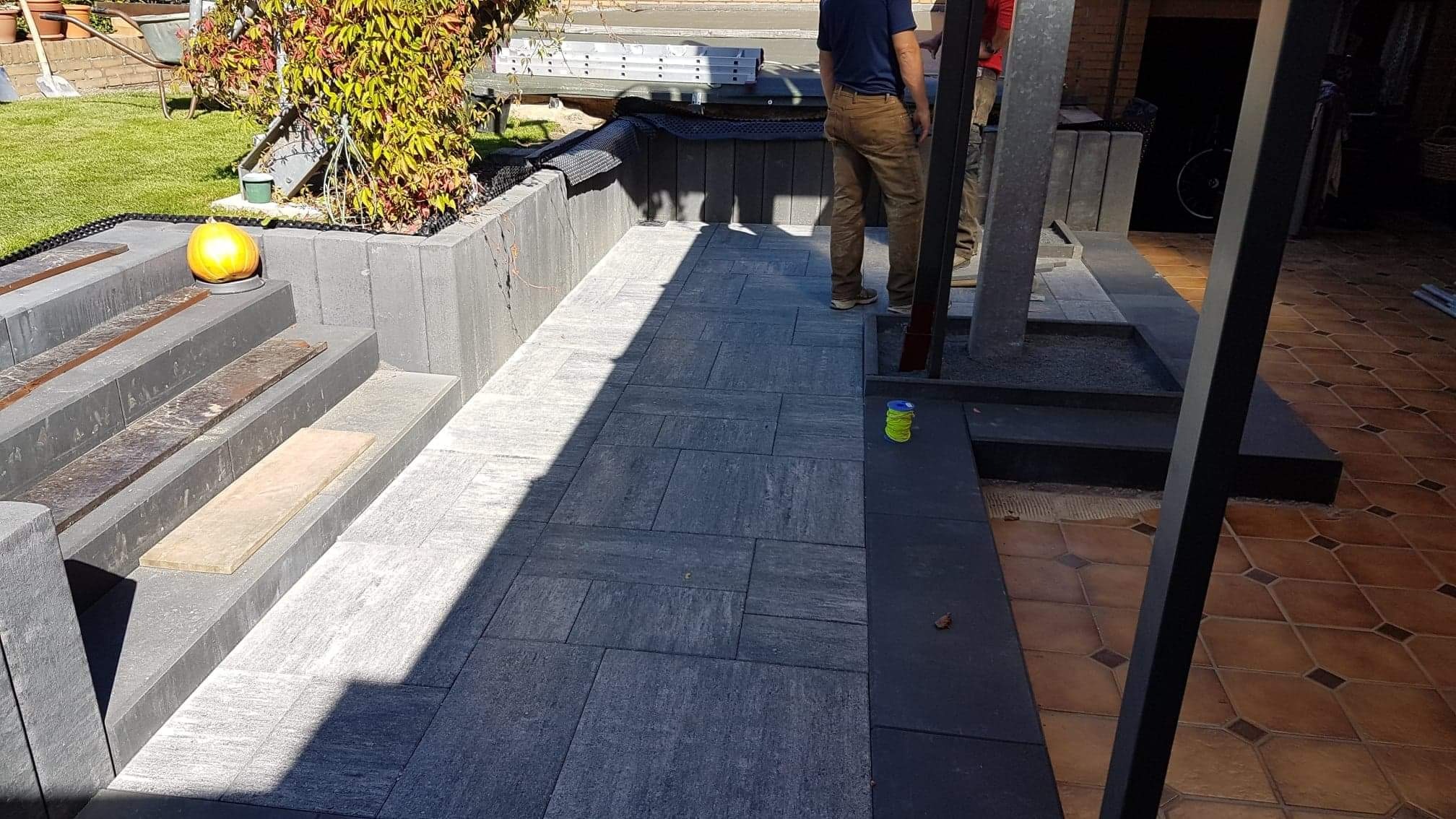 Świeżo ułożona, szara kostka brukowa na posesji, z widocznymi schodami i elementami dekoracyjnymi, takimi jak dynia i zwój sznurka, w tle pracownicy podczas montażu.