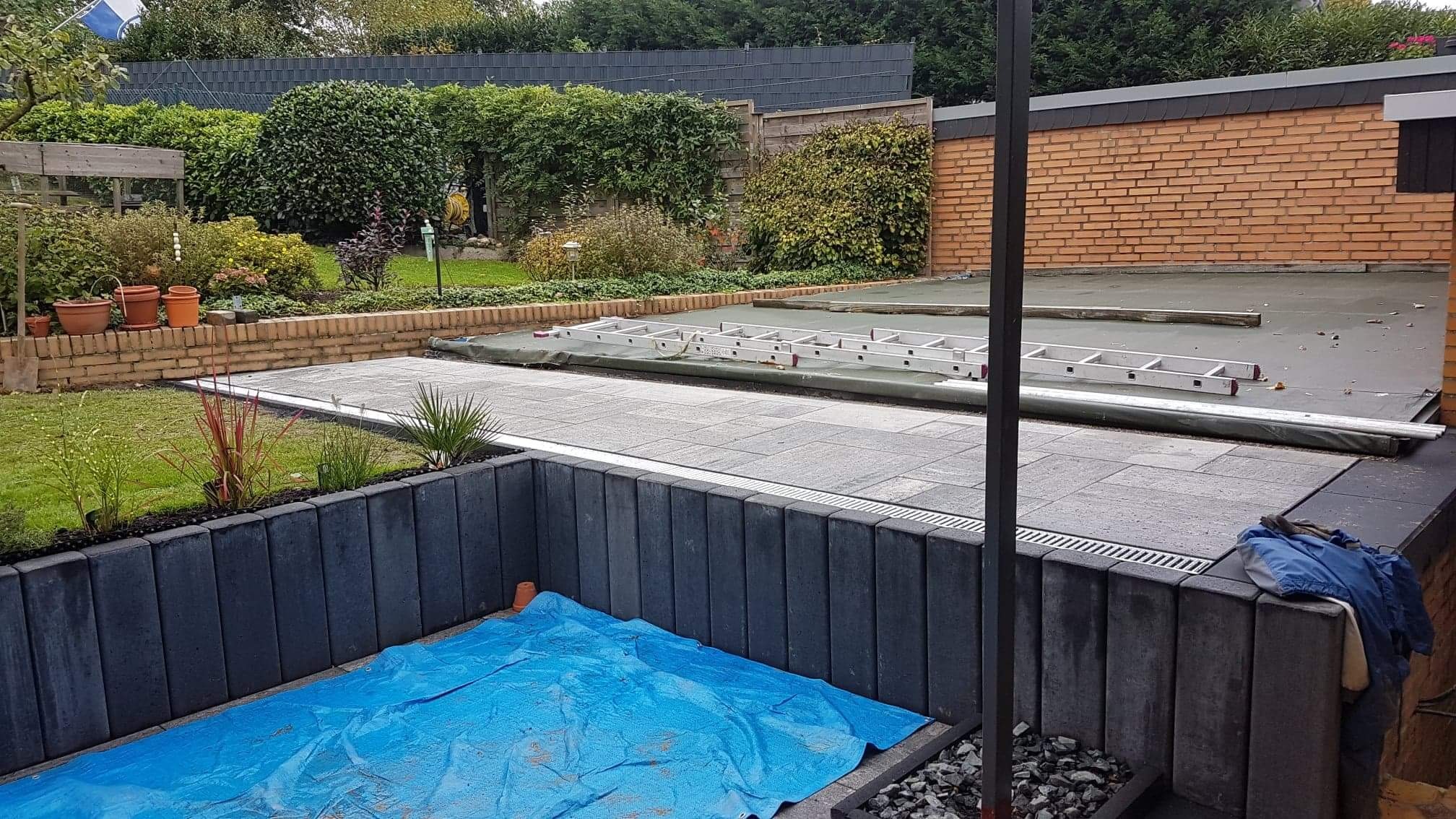 Taras z szarych płyt betonowych, częściowo przykryty plandeką, z widoczną drabinką aluminiową i fragmentem ogrodu z ceglanym obrzeżem.