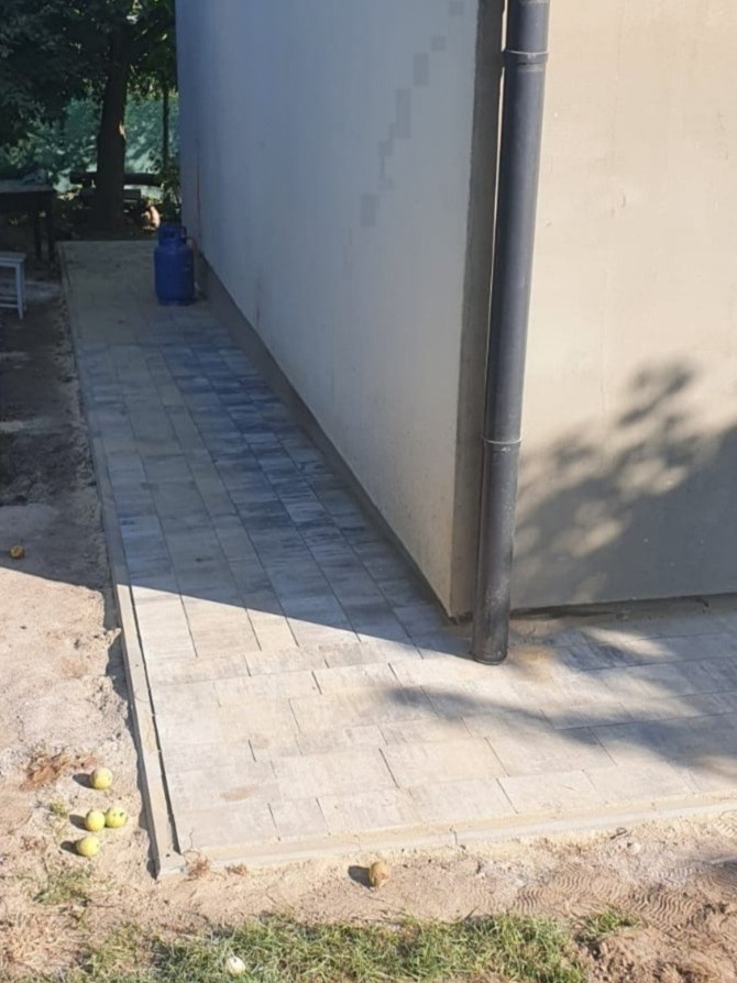 Świeżo ułożona szara kostka brukowa przy ścianie budynku, z widoczną rurą spustową i porozrzucanymi jabłkami na ziemi.