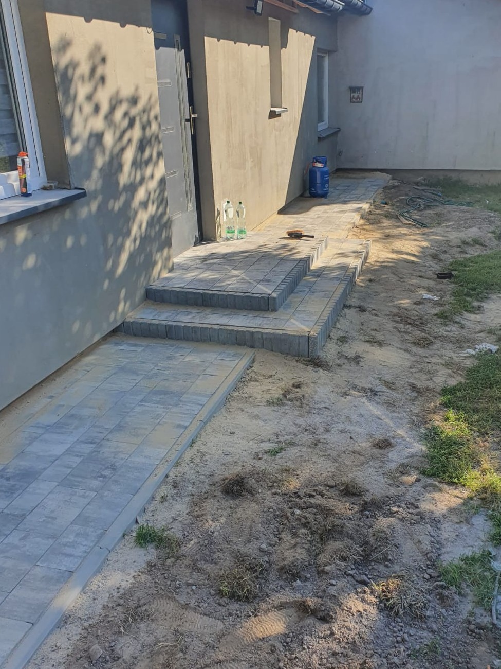 Ułożona kostka brukowa przed wejściem do domu, z trzema stopniami i ścieżką, widoczne butle z gazem i narzędzie brukarskie na schodach.