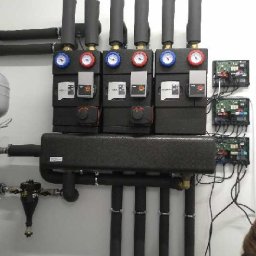 Kus-an Salon Firmowy Viessmann - Rząd czarnych, izolowanych rozdzielaczy z manometrami i termometrami, zamontowanych na białej ścianie, z widocznymi przewodami i elektroniką sterującą.