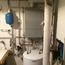 Szary piec wiszący z niebieskim zbiornikiem wyrównawczym i białym zasobnikiem ciepłej wody w piwnicy, widoczne rury i instalacje, obok rury kanalizacyjnej owiniętej izolacją.