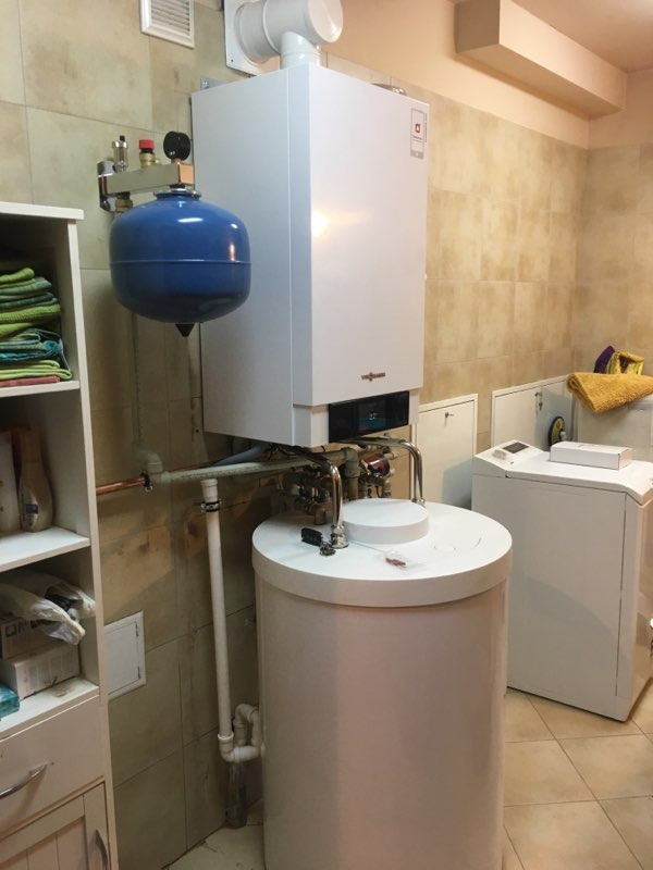 Biały piec gazowy marki Viessmann zamontowany na ścianie w łazience z beżowymi płytkami, z podłączonym niebieskim zbiornikiem wyrównawczym i białym zasobnikiem ciepłej wody; w tle pralka.