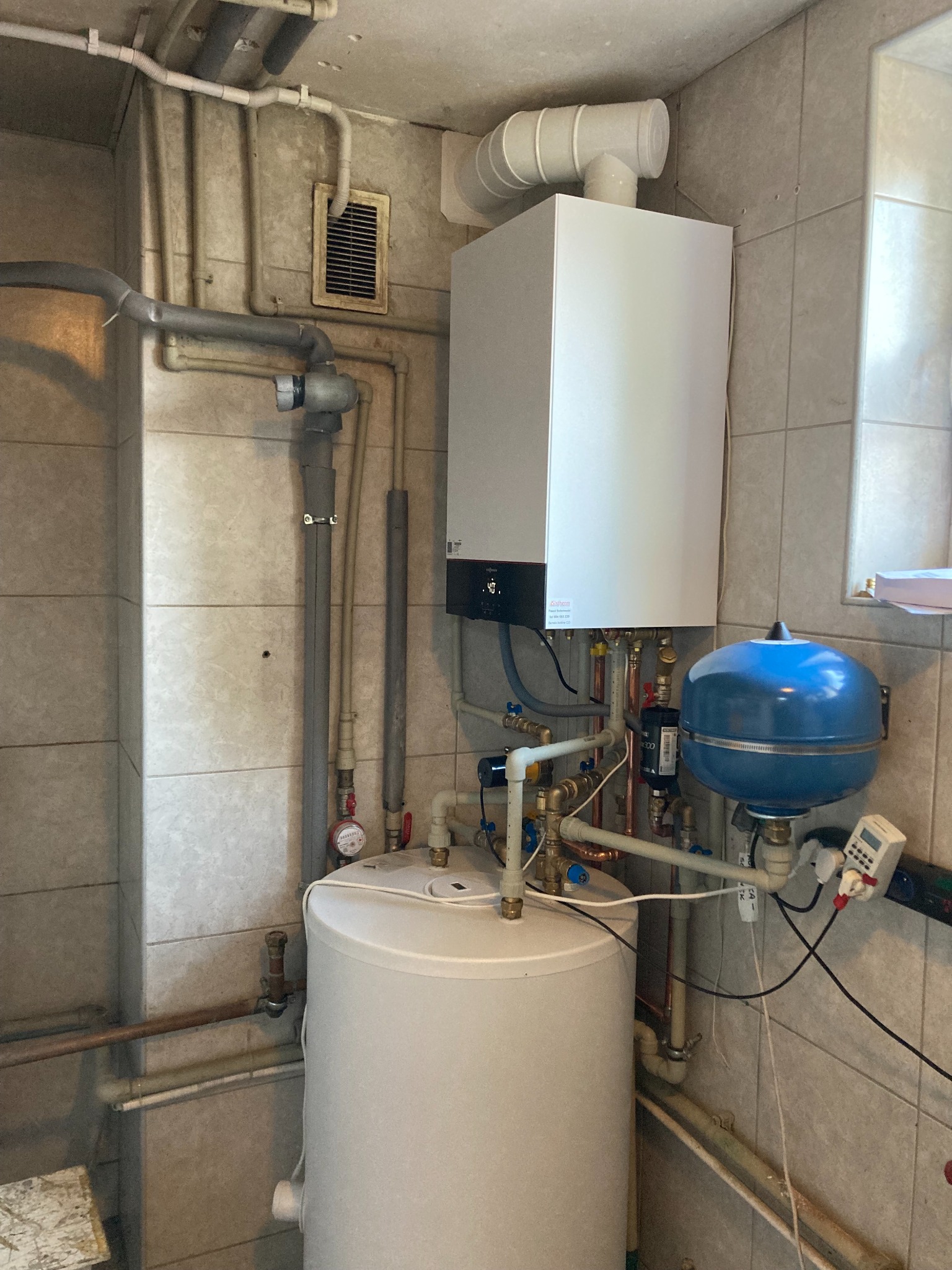 Nowoczesny, biały piec gazowy z niebieskim zbiornikiem wyrównawczym, zamontowany na tle kafelkowanej ściany, widoczne liczne rury i zawory.