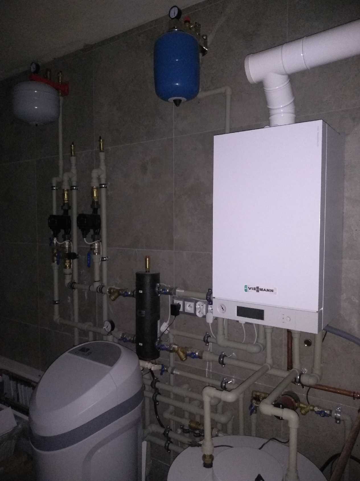 Biały piec gazowy Viessmann z podłączonymi rurami, zbiornikami wyrównawczymi i filtrem wody, zamontowany na tle szarej, kafelkowanej ściany.
