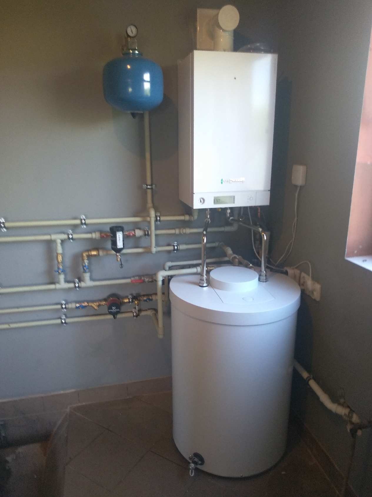 Biały kondensacyjny kocioł gazowy z podłączonym zasobnikiem ciepłej wody i instalacją hydrauliczną na szarej ścianie.