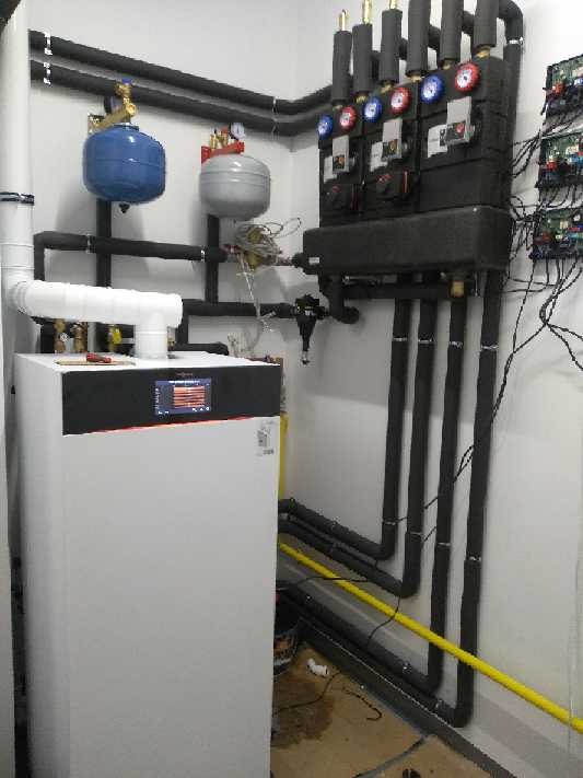 Nowoczesny, biały kocioł z czarnym panelem sterowania i rurami w pomieszczeniu technicznym, widoczne zbiorniki wyrównawcze i rozdzielacz z manometrami.