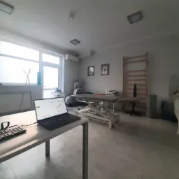 Jasne wnętrze gabinetu z kozetką rehabilitacyjną, drabinką gimnastyczną i biurkiem z laptopem, widok z perspektywy wejścia.