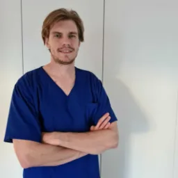 Uśmiechnięty mężczyzna w niebieskim uniformie medycznym, z tatuażem na szyi, stoi na tle białej ściany z założonymi rękami.