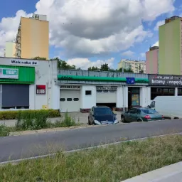 Fasada budynku z napisem 'AWUS Wymiana Opon Mechanika Pojazdowa' z zaparkowanymi samochodami, w tle bloki mieszkalne i pochmurne niebo.