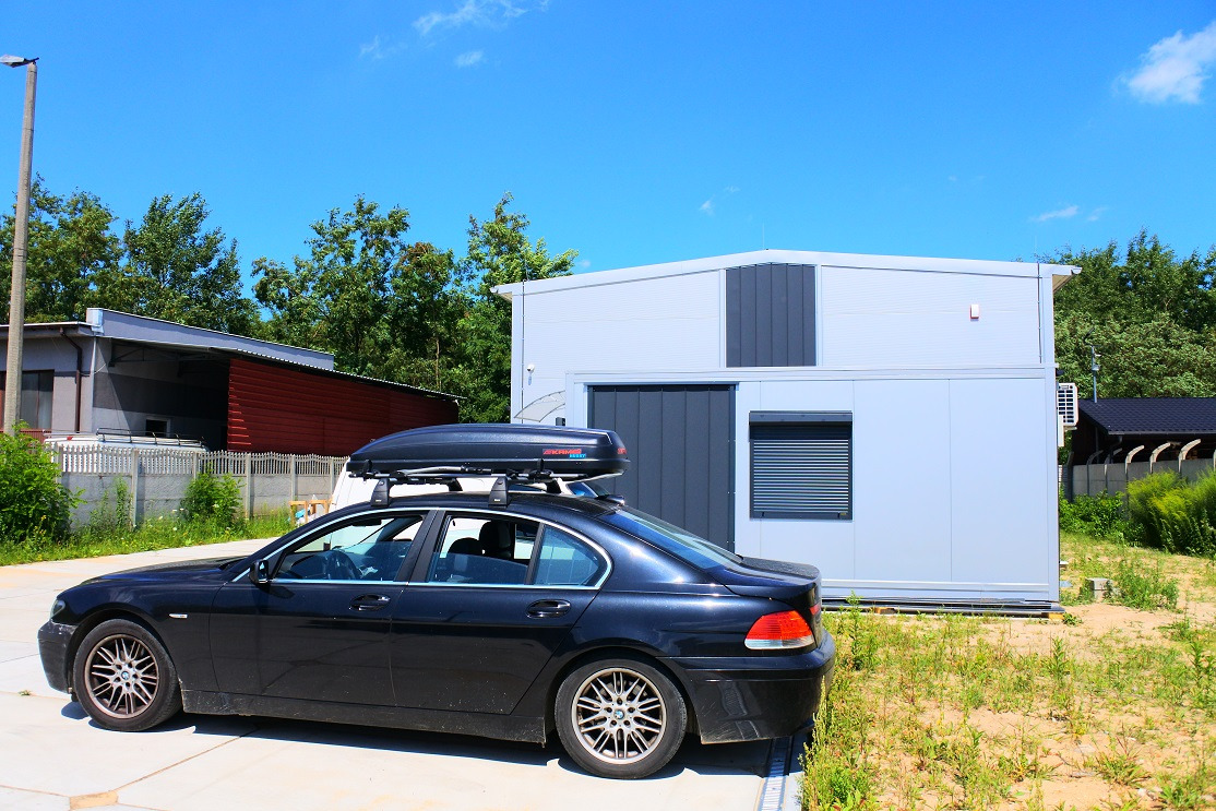 Czarny samochód BMW z bagażnikiem dachowym na tle szarego, modułowego budynku z roletami okiennymi i bramą garażową, w tle inne budynki i zieleń.