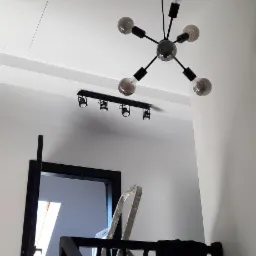 Nowoczesna lampa sufitowa w kształcie gwiazdy z żarówkami dekoracyjnymi, widoczna na tle białego sufitu z dodatkowym oświetleniem punktowym, fragment czarnej balustrady schodów i drabina malarska