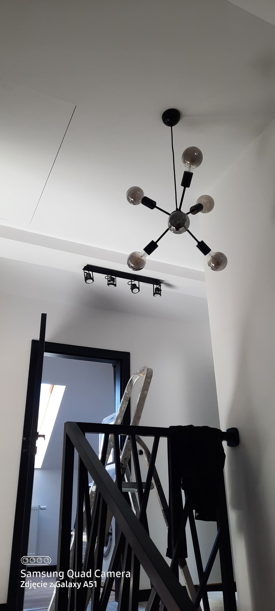 Nowoczesna lampa sufitowa w kształcie gwiazdy z żarówkami dekoracyjnymi, widoczna na tle białego sufitu z dodatkowym oświetleniem punktowym, fragment czarnej balustrady schodów i drabina malarska