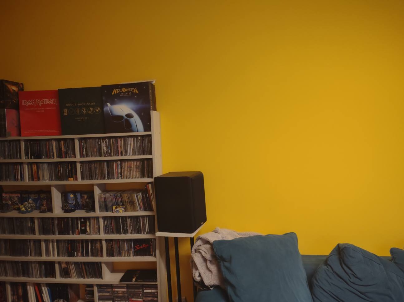 Żółta ściana w pokoju z regałem na płyty CD, na którym stoją płyty Iron Maiden, Bruce Dickinson i Helloween, obok stoi czarny głośnik na białym stojaku, a przed nimi znajduje się kanapa...