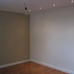 Pomieszczenie po malowaniu: jedna ściana w kolorze taupe, druga biała, drewniana podłoga, sufit z oświetleniem punktowym i wiszącą żarówką bez klosza.