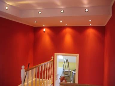 Czerwona ściana świeżo po malowaniu, widoczne halogenowe oświetlenie sufitowe, fragment schodów z drewnianą balustradą i drabina malarska w przejściu do kolejnego pomieszczenia.