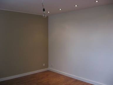 Pomieszczenie po malowaniu: jedna ściana w kolorze taupe, druga biała, drewniana podłoga, sufit z oświetleniem punktowym i wiszącą żarówką bez klosza.