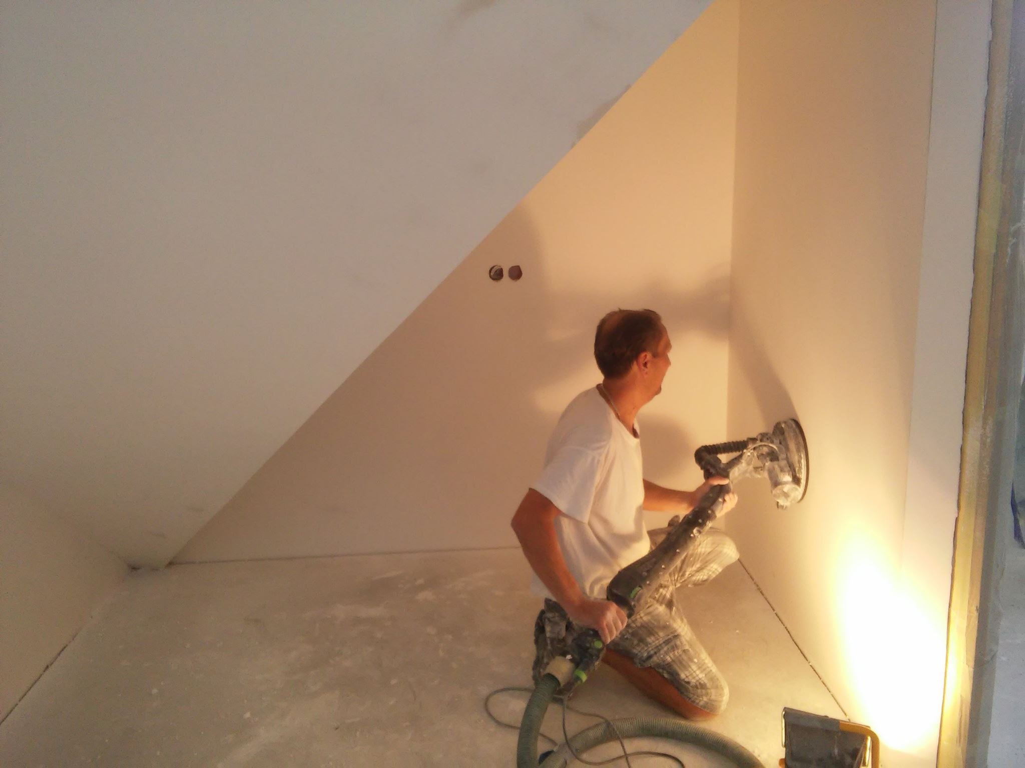 Pracownik szlifuje ściany gładzią za pomocą szlifierki z odkurzaczem w rogu pokoju, widoczne dwa otwory na gniazdka elektryczne w ścianie.