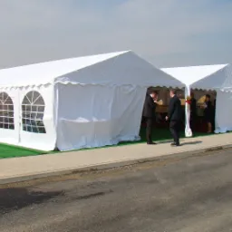 Dwa białe namioty z oknami i falbankami, ustawione na sztucznej trawie, z widocznymi osobami wewnątrz i przed namiotem, oraz flagami reklamowymi w tle.