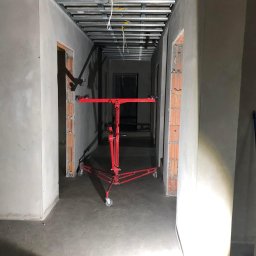 Regips Projekt - Korytarz w trakcie prac wykończeniowych; widoczny metalowy stelaż sufitu podwieszanego, surowe ściany, podłoga wylewka oraz czerwony podnośnik do płyt gipsowo-kartonowych.