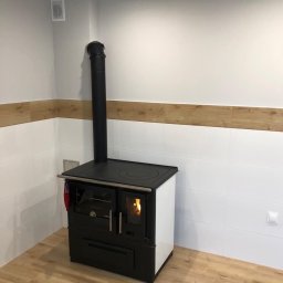 Regips Projekt - Kuchenka kaflowa z czarnym kominem w rogu pomieszczenia z białymi kaflami i drewnopodobną listwą, płomienie widoczne przez szybę.