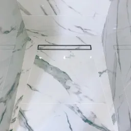 Kabina prysznicowa wykończona białymi płytkami imitującymi marmur z odpływem liniowym.