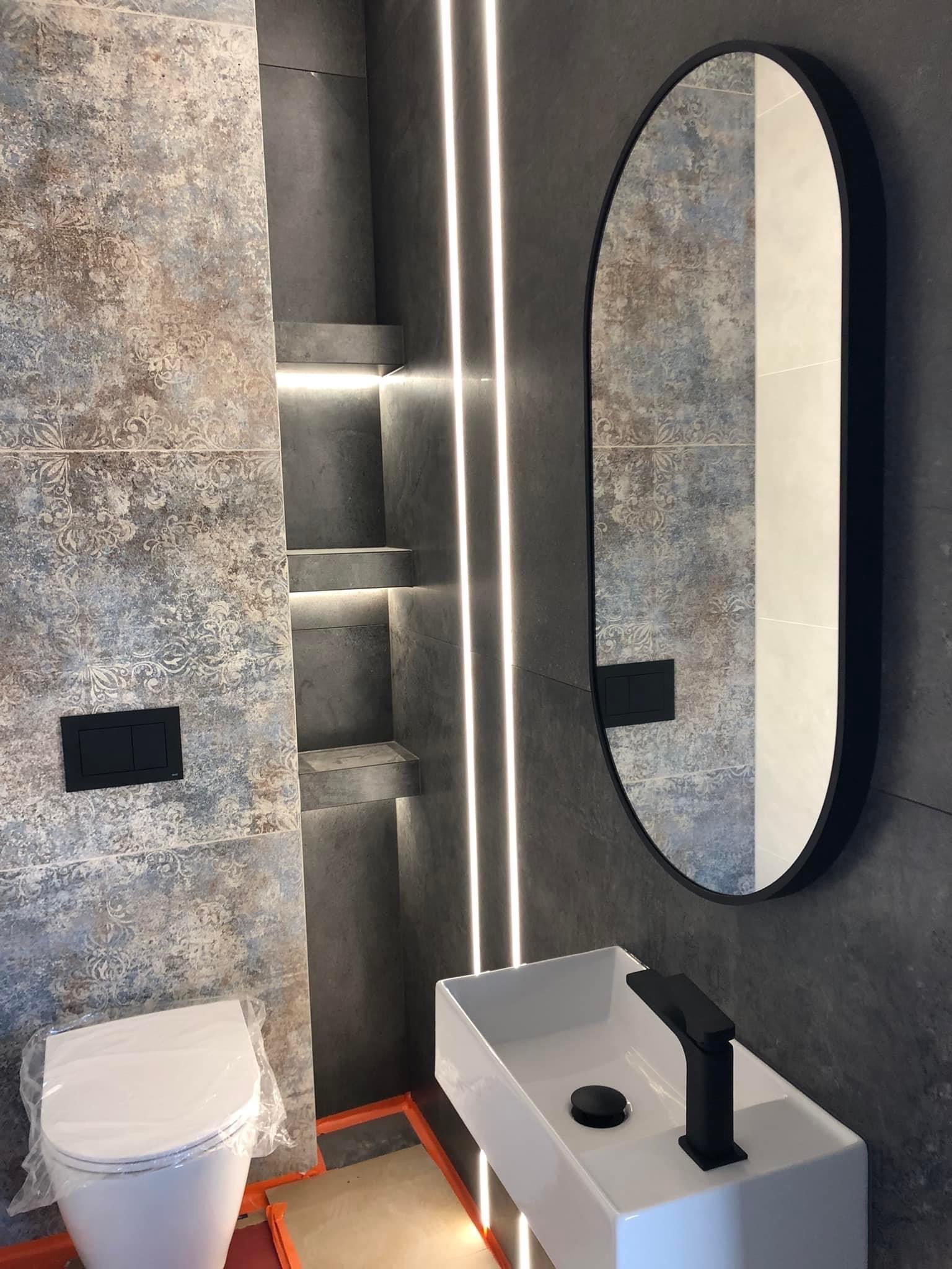 Nowoczesna łazienka z szaro-niebieskimi płytkami w stylu vintage, podświetlane półki w ścianie, owalne lustro w czarnej ramie i biała umywalka z czarną armaturą. Toaleta zabezpieczona folią ochronną.