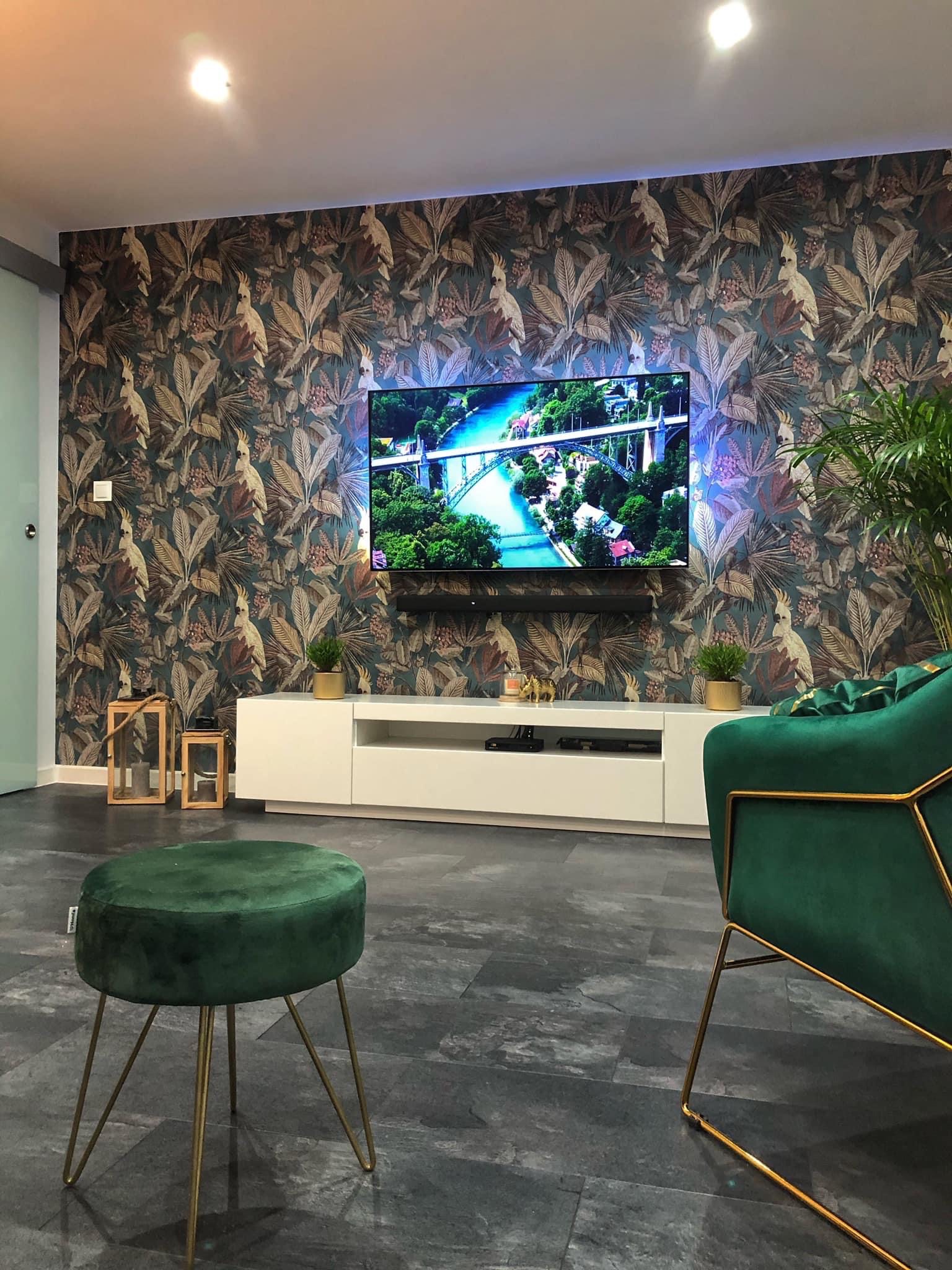 Salon z tapetą w tropikalne wzory, telewizorem podświetlonym na niebiesko, białą szafką RTV, zielonym pufem na złotych nóżkach i zielonym fotelem.