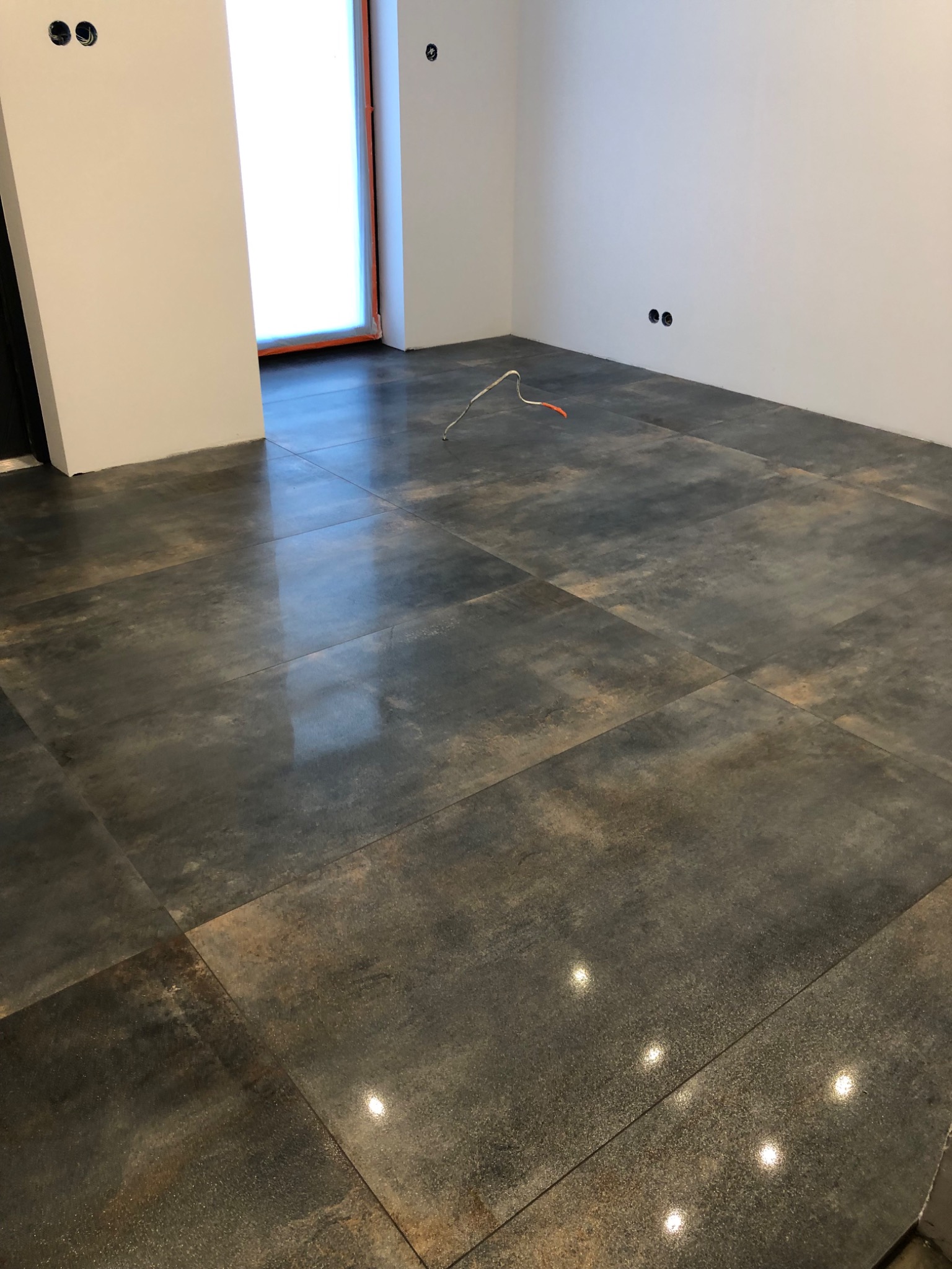 Ułożona podłoga z dużych, ciemnych płytek imitujących metal, odbijających światło, z widocznymi punktami elektrycznymi w ścianie i wystającymi kablami.
