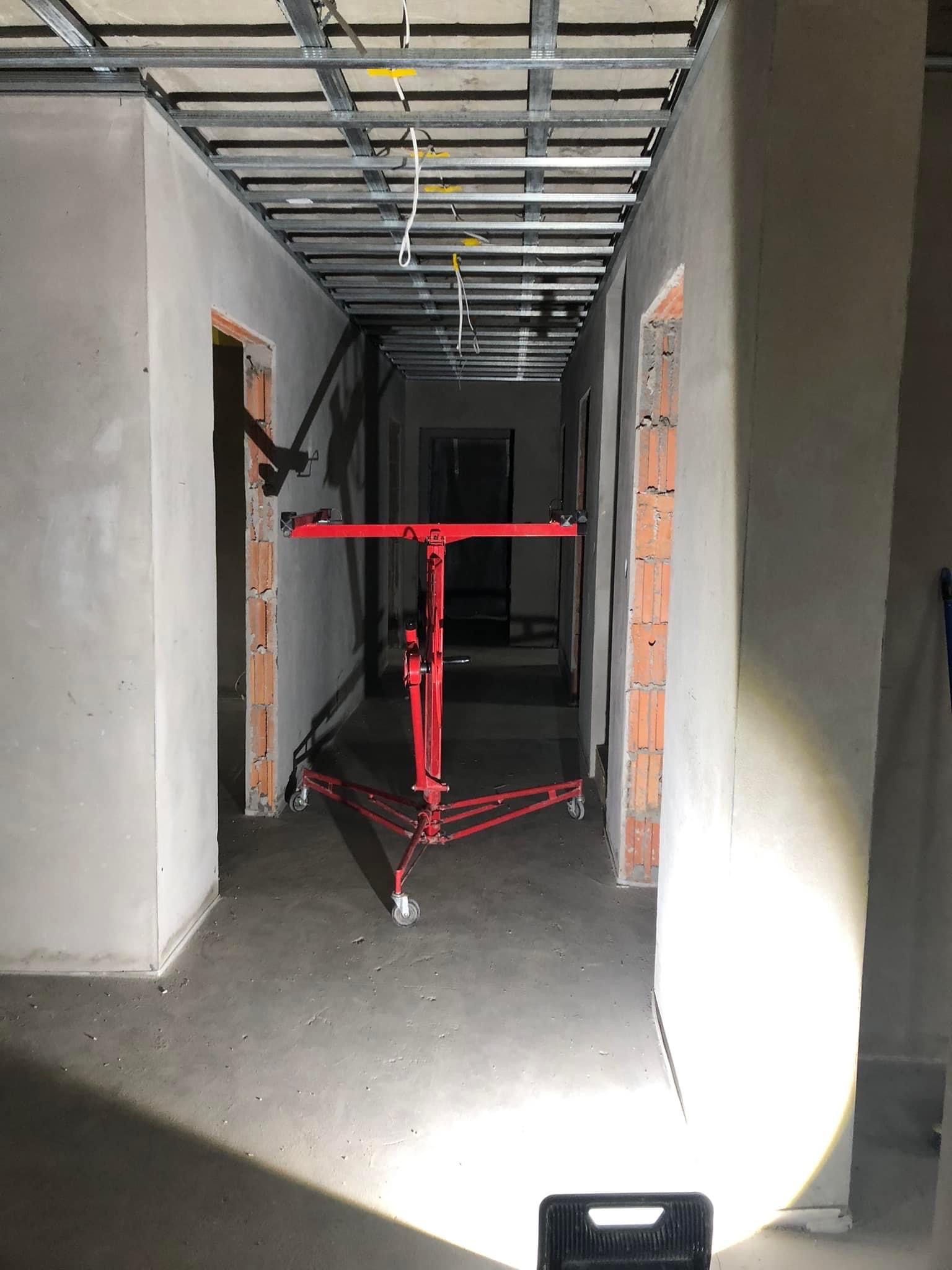 Korytarz w trakcie prac wykończeniowych; widoczny metalowy stelaż sufitu podwieszanego, surowe ściany, podłoga wylewka oraz czerwony podnośnik do płyt gipsowo-kartonowych.