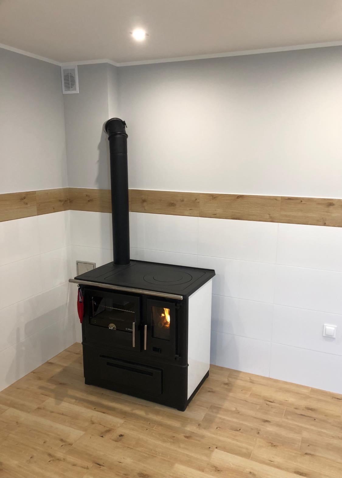 Kuchenka kaflowa z czarnym kominem w rogu pomieszczenia z białymi kaflami i drewnopodobną listwą, płomienie widoczne przez szybę.