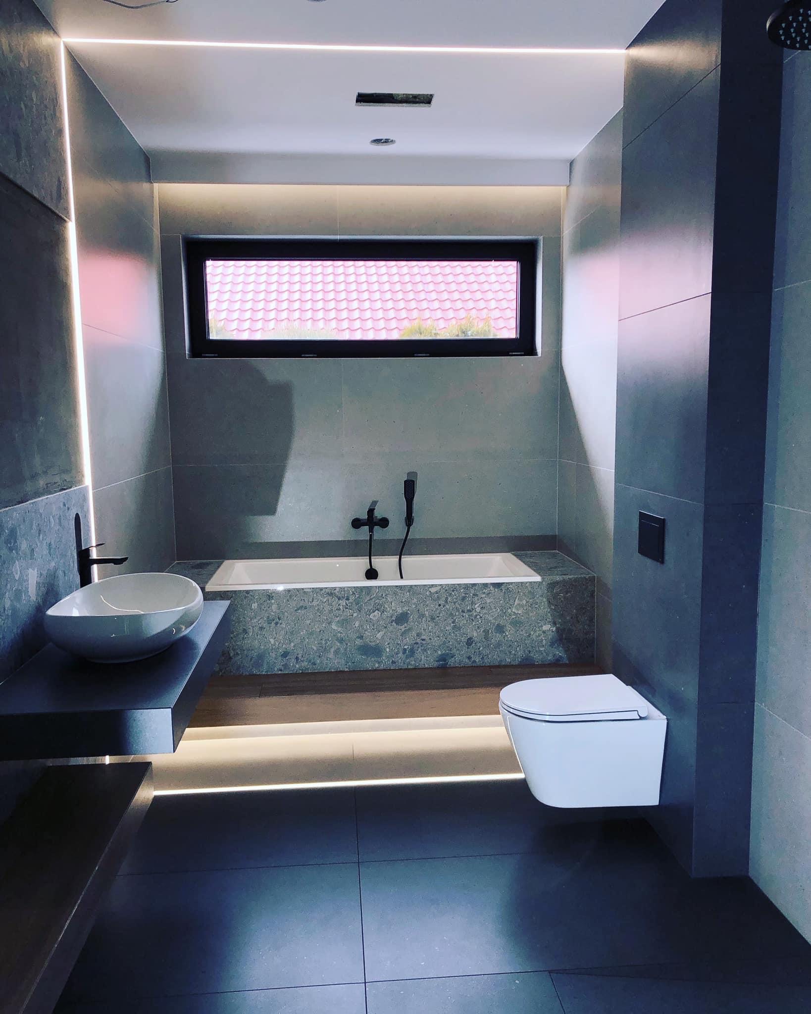 Nowoczesna łazienka w szarościach z podświetleniem LED, owalna umywalka nablatowa, wisząca toaleta i wanna z obudową z kamienia, okno z widokiem na dachówkę.