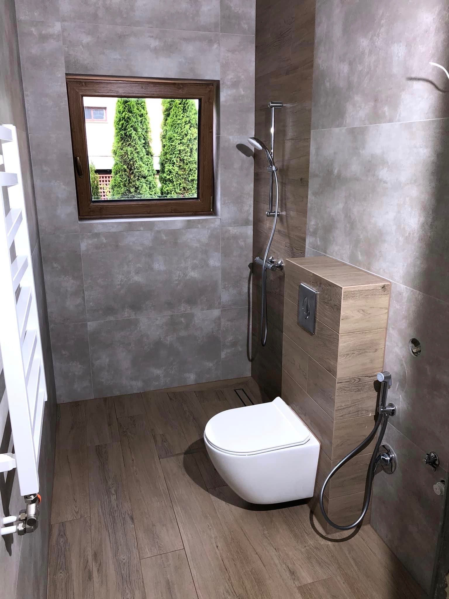 Nowoczesna łazienka z szarymi płytkami imitującymi beton, drewnianą podłogą i oknem z widokiem na tuje.
