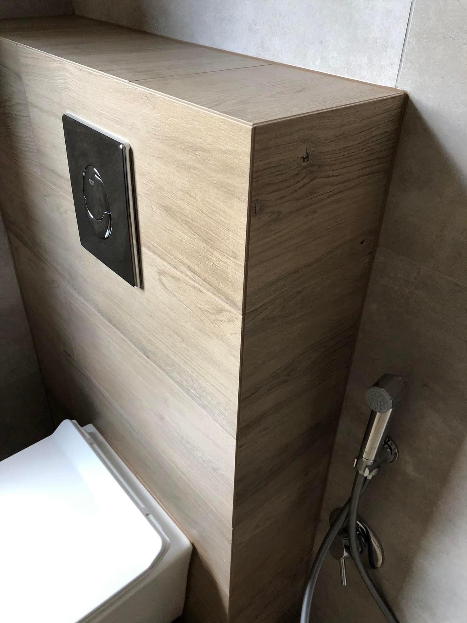 Podtynkowa zabudowa toalety z płytkami imitującymi drewno, chromowany przycisk spłukiwania i bidetka natryskowa.