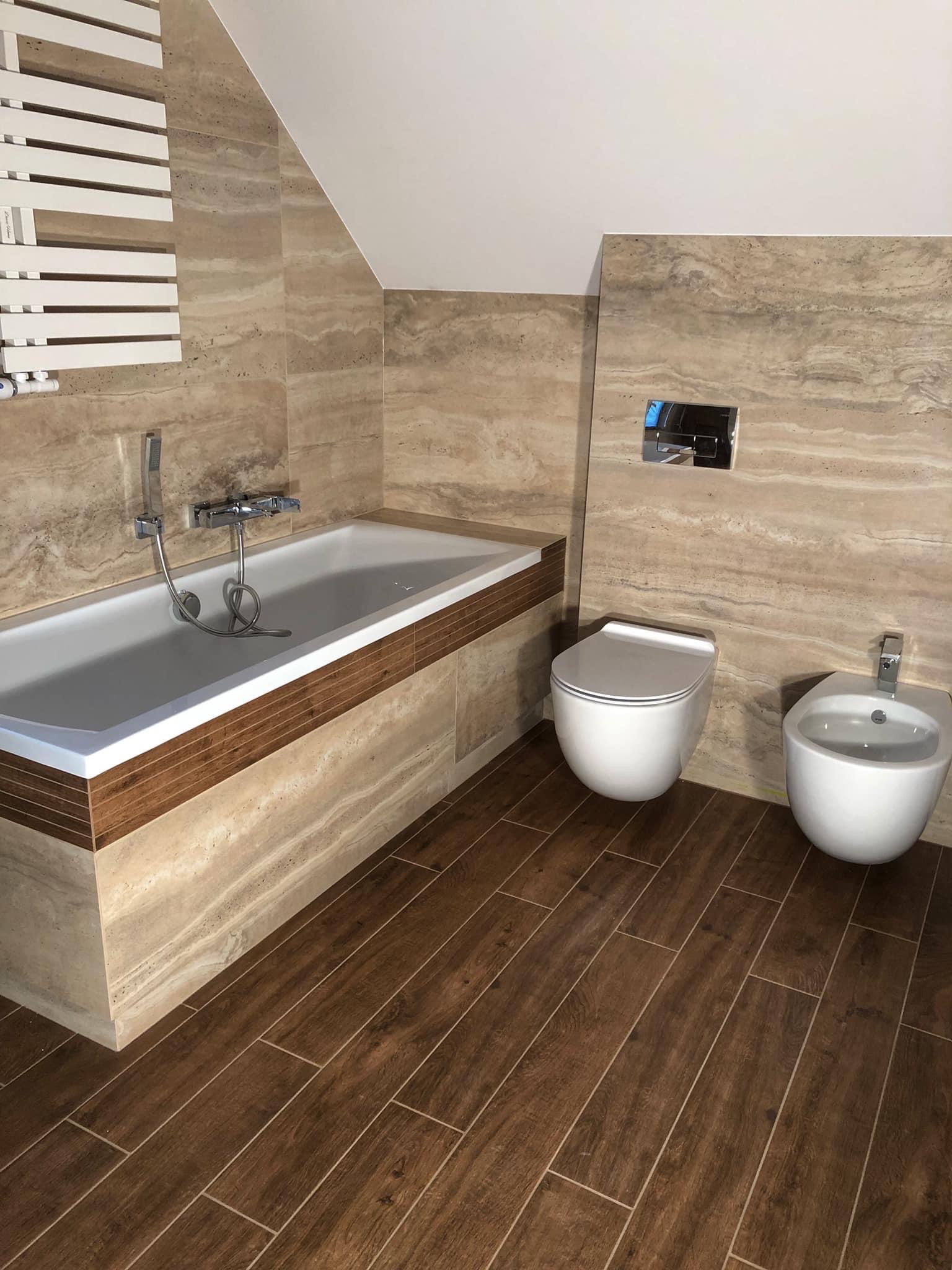 Nowoczesna łazienka na poddaszu z beżowymi płytkami imitującymi marmur na ścianach i drewnopodobną podłogą, biała wanna z chromowaną armaturą, wisząca toaleta i bidet.