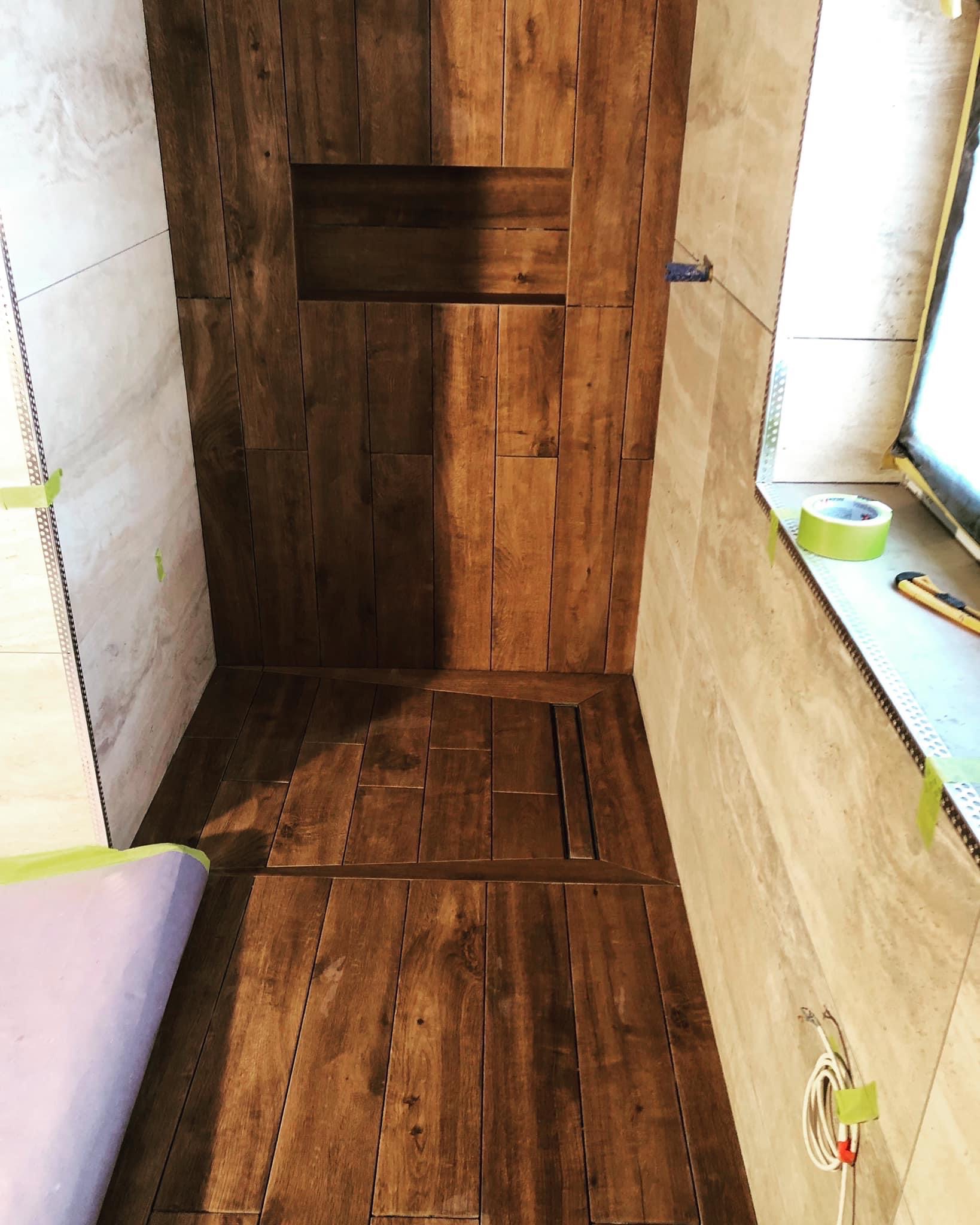 Wnętrze kabiny prysznicowej z wykończeniem imitującym drewno, widoczny odpływ liniowy i wnęka w ścianie, jasne płytki na ścianach, zabezpieczone taśmą malarską.