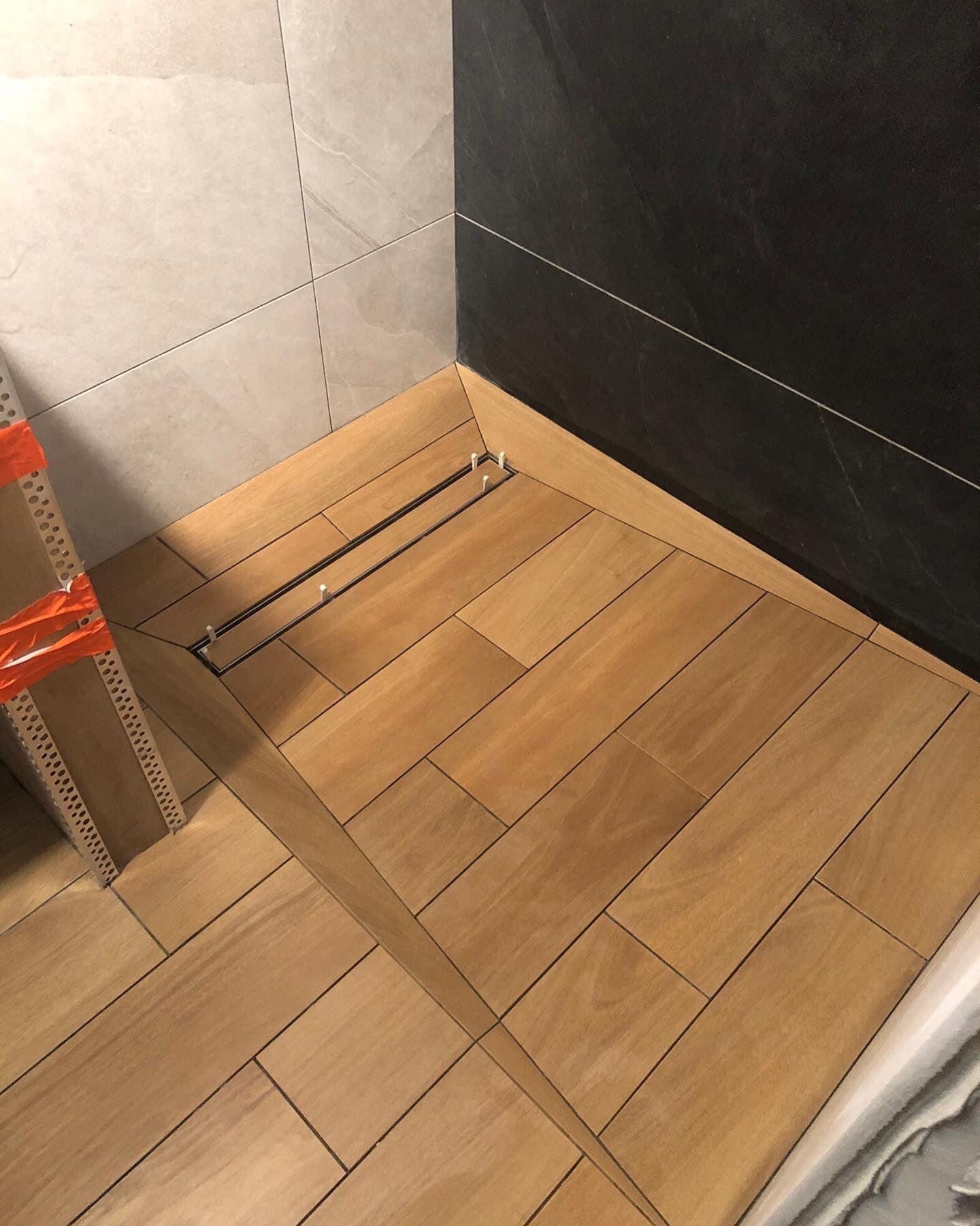 Ułożone drewnopodobne płytki podłogowe w kabinie prysznicowej z odpływem liniowym, widoczne profile narożnikowe zabezpieczone pomarańczową taśmą.