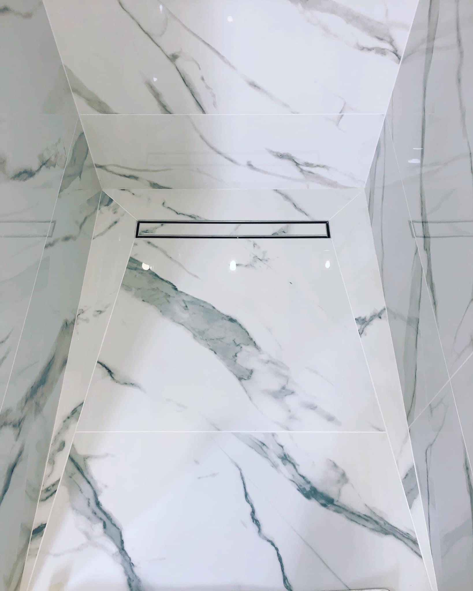 Kabina prysznicowa wykończona białymi płytkami imitującymi marmur z odpływem liniowym.