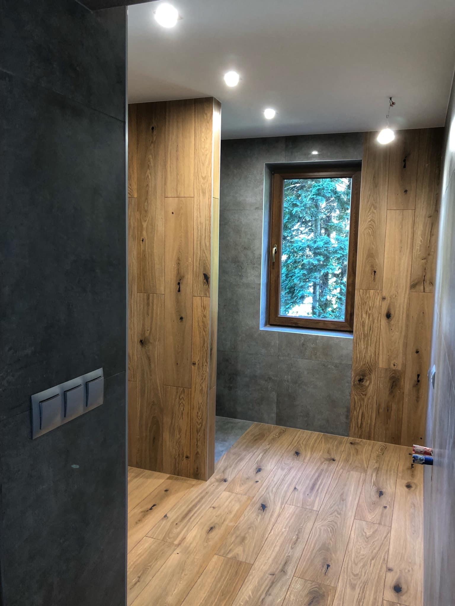 Nowoczesna łazienka z drewnianymi panelami ściennymi i podłogą, szare płytki na ścianach, okno z widokiem na drzewa, minimalistyczne włączniki światła.