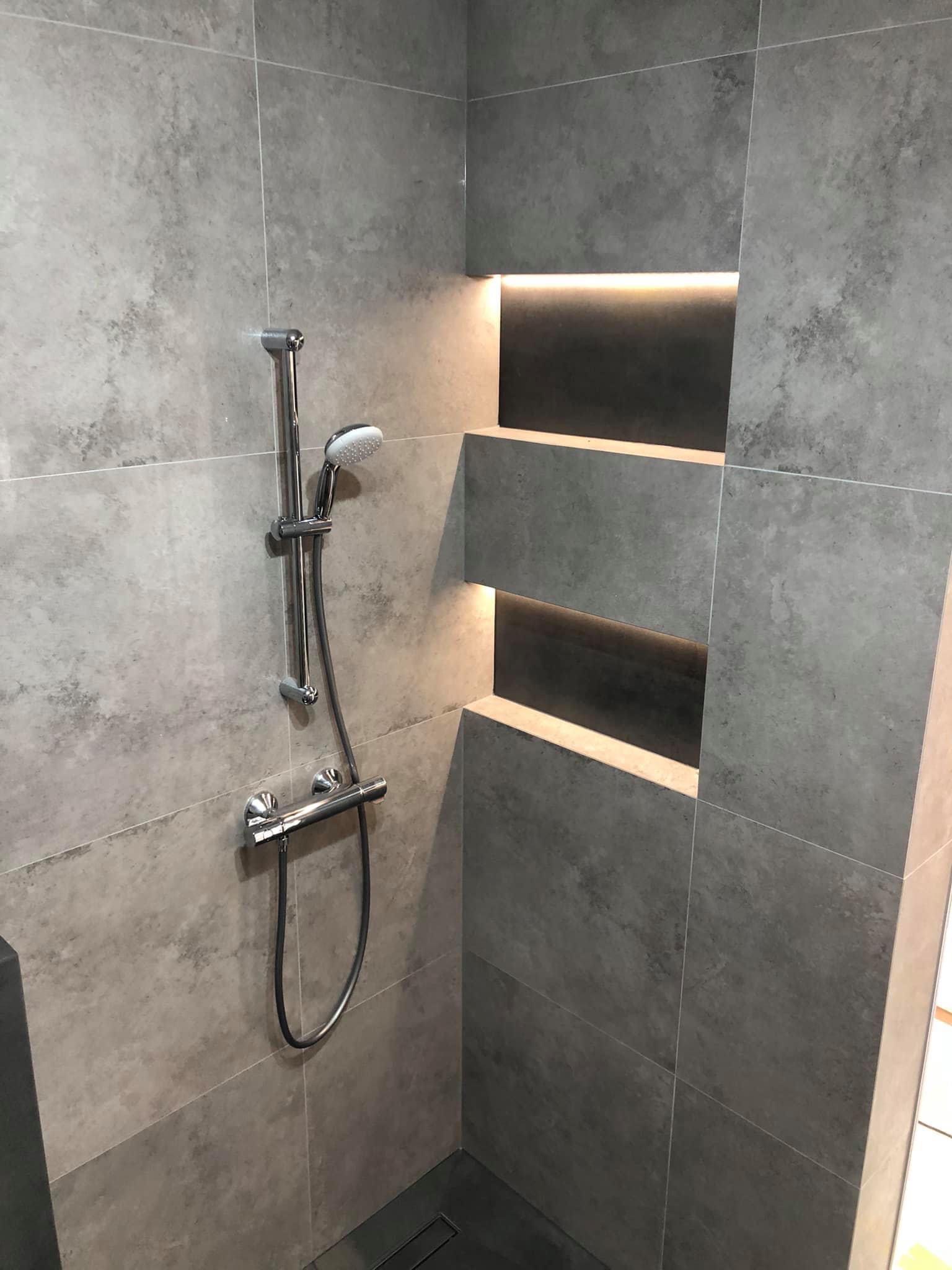 Nowoczesny prysznic z szarymi płytkami imitującymi beton, chromowaną armaturą i podświetlanymi wnękami w ścianie.