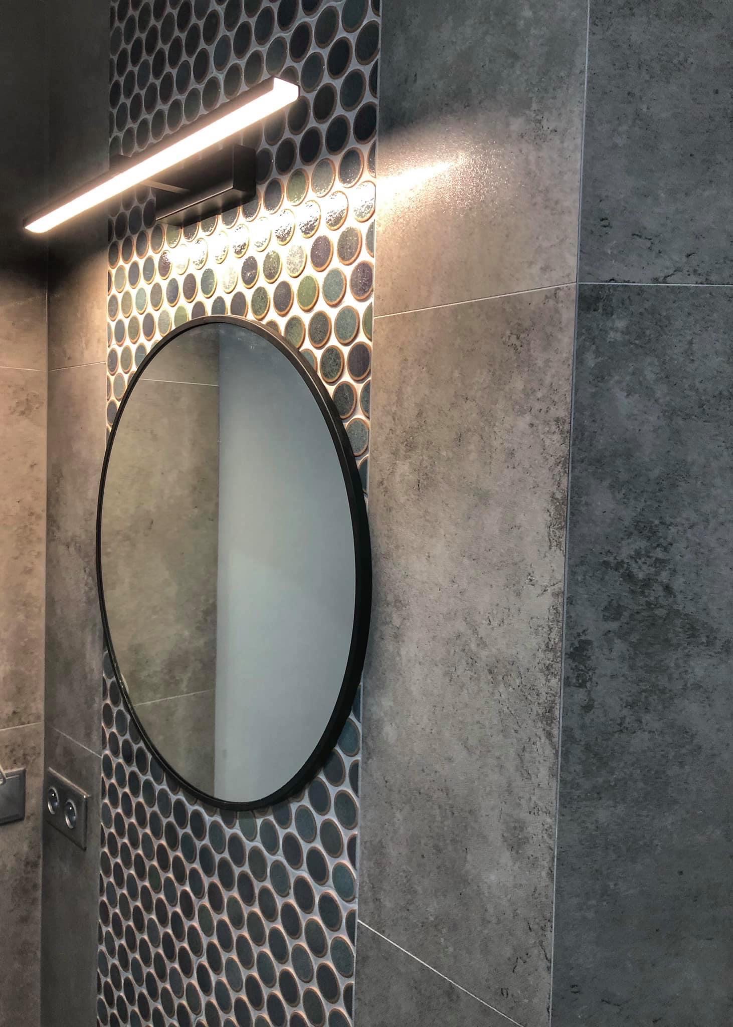 Nowoczesna łazienka z okrągłym lustrem, podświetlaną lampą LED i mozaiką z okrągłych płytek w odcieniach szarości i zieleni, uzupełniona szarymi płytkami wielkoformatowymi.