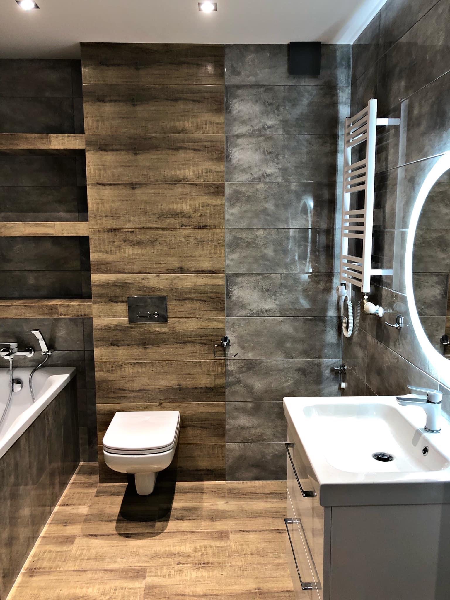Nowoczesna łazienka z drewnopodobnymi i szarymi płytkami, podświetlanym lustrem, grzejnikiem drabinkowym i wiszącą toaletą.