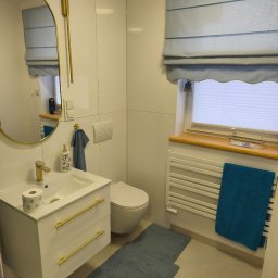 MAT-BUD MATEUSZ MASNY - Biała łazienka z armaturą w kolorze złota: umywalka z szafką, toaleta, lustro i grzejnik. Błękitne akcenty: roleta rzymska, ręczniki i dywaniki.