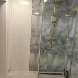 MAT-BUD MATEUSZ MASNY - Nowoczesna kabina prysznicowa z deszczownicą w złotej tonacji, wykończona płytkami imitującymi marmur. Białe ściany i podłoga podkreślają minimalistyczny design łazienki.