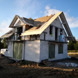 Surowy stan domu z białych bloczków, widoczne drewniane elementy konstrukcji dachu, okna zabezpieczone folią, na zewnątrz ziemia i materiały budowlane.