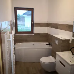 Nowoczesna łazienka po remoncie w Kościerzynie: biała wanna narożna, podwieszana toaleta i umywalka z szafką, brązowo-białe płytki na ścianach, widok z okna na ogród.