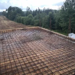 Zbrojenie płyty fundamentowej na deskowaniu, widok z góry, w tle łąka i las, niebo pochmurne.