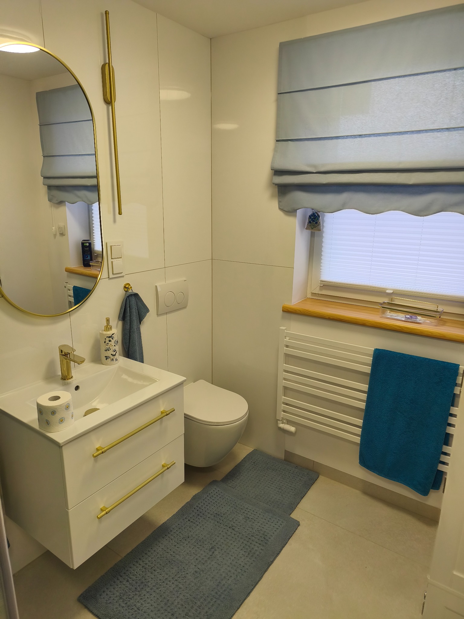 Biała łazienka z armaturą w kolorze złota: umywalka z szafką, toaleta, lustro i grzejnik. Błękitne akcenty: roleta rzymska, ręczniki i dywaniki.