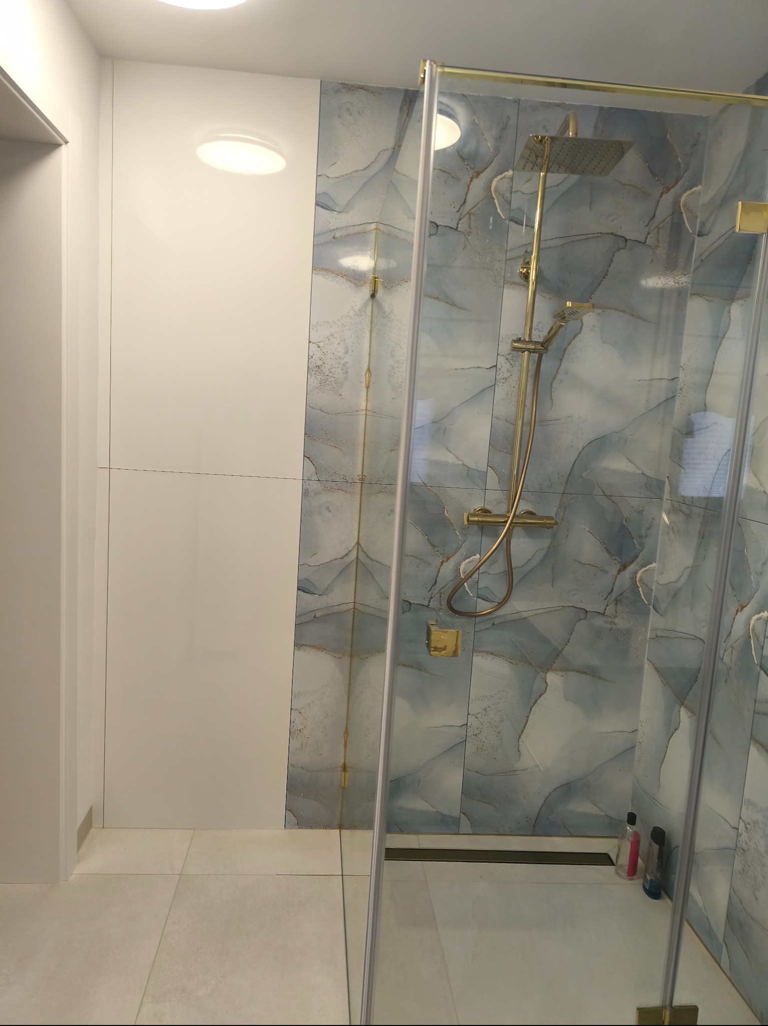 Nowoczesna kabina prysznicowa z deszczownicą w złotej tonacji, wykończona płytkami imitującymi marmur. Białe ściany i podłoga podkreślają minimalistyczny design łazienki.