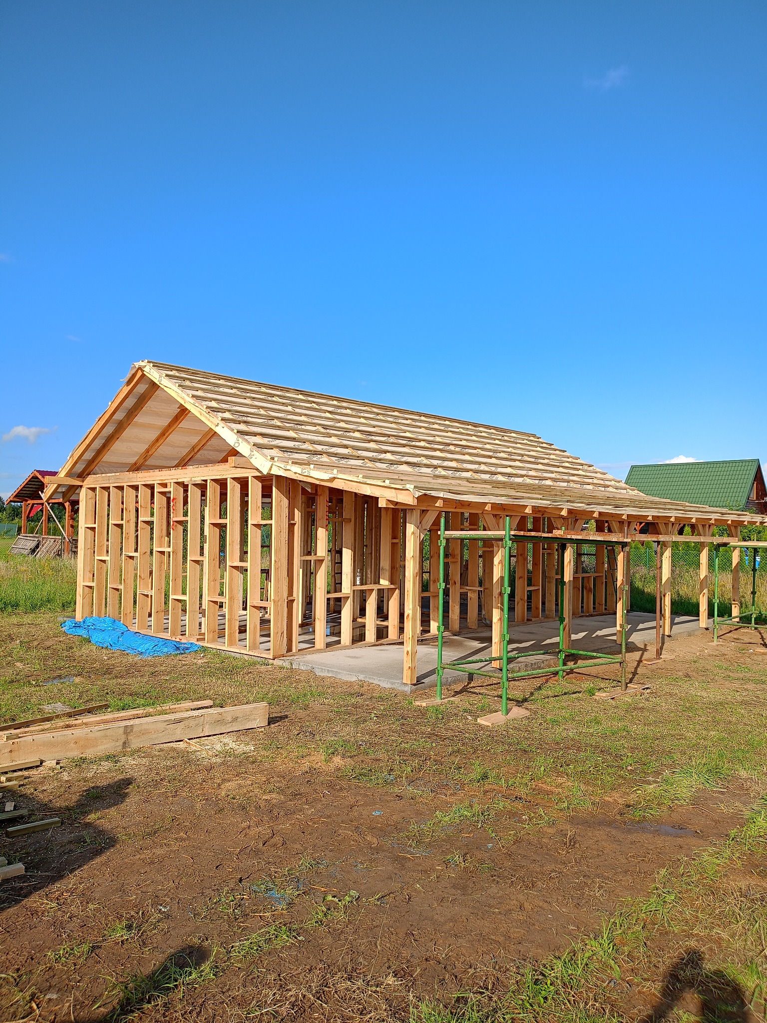 Konstrukcja szkieletowa domu w trakcie budowy, widoczne drewniane elementy nośne i zadaszenie. W tle błękitne niebo i zieleń. Częściowo widoczne rusztowania.